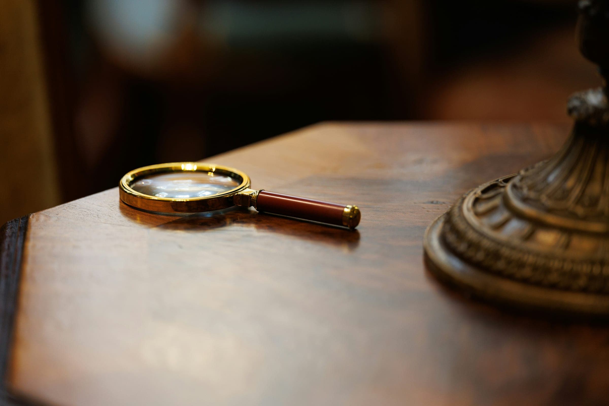 A brass magnifier resting on a polished wooden desk beside an ornate bronze pedestal.